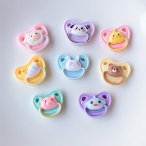 May include: A collection of colorful baby pacifiers in various shapes and designs. Each pacifier features a different animal or character, including a cow, dog, chick, bunny, bear, pig, and dolphin. The pacifiers are in shades of pink, yellow, blue, and purple.