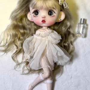 May include: A doll with long, wavy blonde hair and large, expressive eyes. The doll wears a white dress, white stockings, and a bow-shaped accessory on its head. A small, clear bottle is next to the doll.