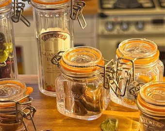 Puede incluir: Varios tarros de vidrio transparente con sellos de goma naranja y cierres metálicos. Los tarros son de diferentes formas y tamaños, algunos contienen especias. Se puede ver el nombre de la marca "Kilner".