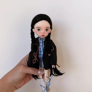 Può includere: Una bambola snodabile con lunghi capelli neri raccolti in due trecce, che indossa un blazer nero gessato sopra una camicia a quadri blu e bianchi, una gonna grigia, calzini bianchi con lacci a nastro blu e scarpe da ginnastica chiare.