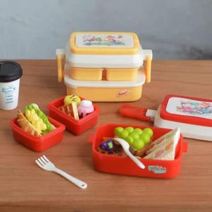 May include: A selection of miniature toy food and lunch boxes. The set includes yellow and white stacked lunch boxes, red containers with various food items, a white fork, and a coffee cup. The lunch boxes have cartoon illustrations on the lids.
