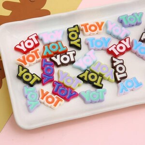 May include: A collection of colorful plastic beads spelling out "TOY" and "YOT" in various colors, including red, orange, blue, and green. The beads are arranged on a white rectangular tray, suitable for crafting or jewelry making.