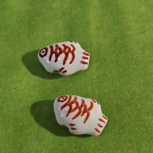 May include: Two small, white ceramic fish figurines with brown details, including eyes, gills, and tail markings. The fish are set against a green background.