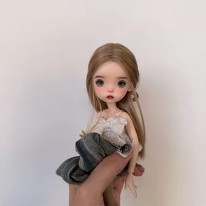 Puede incluir: Una muñeca con pelo largo rubio, ojos azules y mejillas rosadas. La muñeca lleva un top blanco con un lazo y una falda vaquera. La muñeca mide aproximadamente 15 cm de altura.