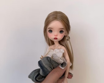 May include: A doll with long blonde hair, blue eyes, and rosy cheeks. The doll wears a white top with a bow and a denim skirt. The doll is approximately 6 inches tall.