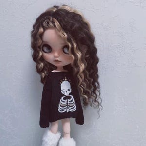 Op de afbeelding: Pop met grote ogen en lang, golvend bruin en blond haar. De pop draagt een zwarte trui met een wit skeletontwerp en pluizige witte laarzen. De achtergrond is lichtgrijs.