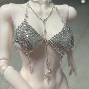 Könnte beinhalten: Ein silbernes Ketten-Bikini-Top und Halsketten-Set. Das Bikini-Top besteht aus kleinen, reflektierenden Metallscheiben, und die Halskette hat eine zarte Kette mit kleinen Perlen und einem Schlangenanhänger. Das Set ist als Modeaccessoire konzipiert.