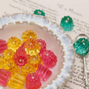 May include: A collection of colorful, translucent, flower-shaped charms in shades of yellow, pink, and green, each with a small, sparkling gem in the center. They are arranged in a decorative, scalloped-edge dish, suggesting a craft or jewelry theme.