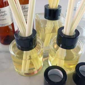 May include: Four clear glass bottles with black lids and white diffuser sticks. The bottles are filled with a light yellow liquid. The bottles are on a white marble surface. The bottles are labelled "Your Crafts" and "Dreams".