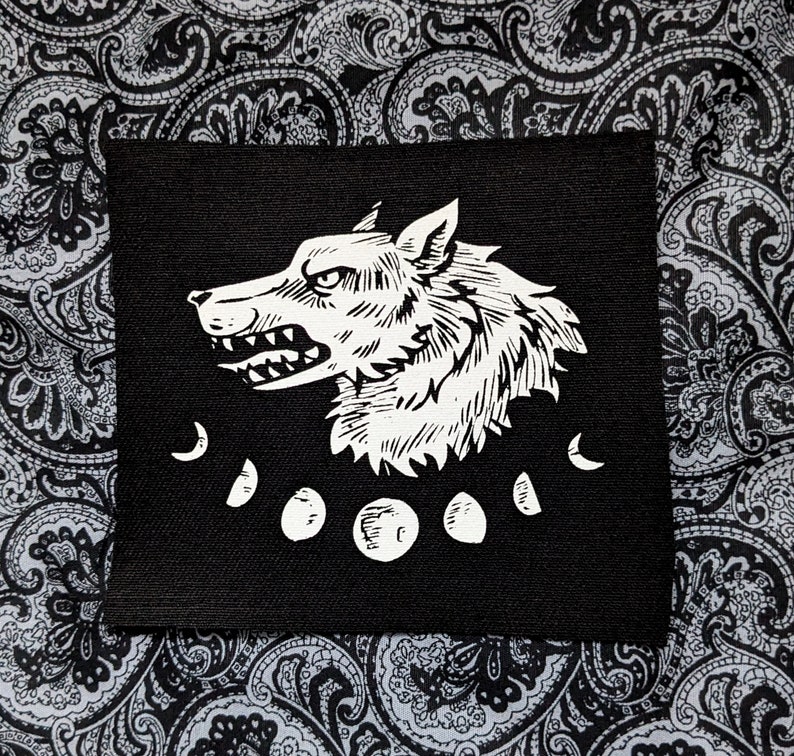 May include: Black and white embroidered patch of a wolf's head with a crescent moon and full moon phases around the neck.