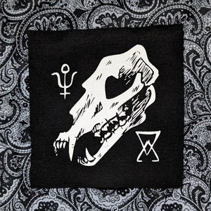 May include: Black and white embroidered patch featuring a skull with sharp teeth, a symbol of the planet Uranus, and a pentagram. The patch is on a black fabric background.