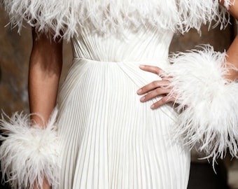 Può includere: Un abito bianco plissettato con top di paillettes e dettagli in piuma. L'abito presenta uno scollo tondo e un corpetto aderente. Le maniche sono decorate con piume bianche e i polsi hanno polsini in piuma abbinati. La modella indossa grandi orecchini a goccia.