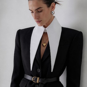 Puede incluir: Un blazer negro con cuello blanco y cinturón negro. El blazer tiene un collar con colgante de corazón dorado y pendientes de aro dorados. El conjunto es de estilo clásico y elegante.