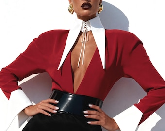 Può includere: Una camicetta rossa con scollatura profonda e colletto bianco, abbinata a pantaloni neri e una cintura nera larga. La camicetta ha polsini bianchi e un dettaglio stringato sul colletto. Orecchini dorati completano il look.