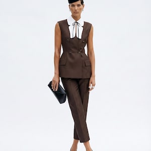 Pu&ograve; includere: Un completo marrone con gilet doppiopetto, pantaloni abbinati e cappello nero. Il completo presenta una camicia bianca con colletto e cravatta nera. L'outfit &egrave; completato da una pochette nera e scarpe nere con punte dorate.
