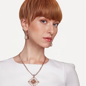May include: A woman wearing a gold necklace with a diamond-shaped pendant, and matching earrings. The pendant features a central design surrounded by white and gold accents. The woman is wearing a white dress.