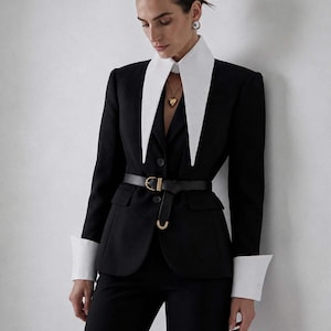 Op de afbeelding: Een zwarte blazer met een witte kraag en manchetten, gecombineerd met een zwarte broek. De blazer heeft een zwarte riem met een gouden gesp. Een gouden hartvormige hanger.