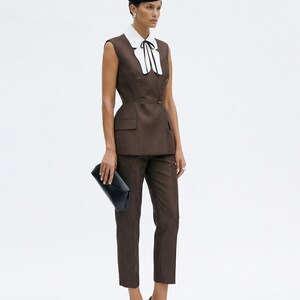 Pu&ograve; includere: Completo marrone composto da un blazer senza maniche con colletto bianco e fiocco nero, abbinato a pantaloni coordinati. L'outfit &egrave; accessoriato con una pochette nera e tacchi neri con punte dorate. Un cappello nero completa l'ensemble.