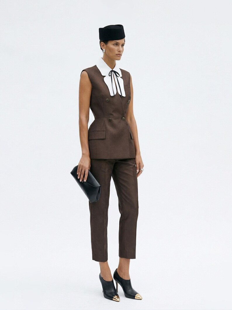 Pu&ograve; includere: Completo marrone composto da un blazer senza maniche con colletto bianco e fiocco nero, abbinato a pantaloni coordinati. L'outfit &egrave; accessoriato con una pochette nera e tacchi neri con punte dorate. Un cappello nero completa l'ensemble.