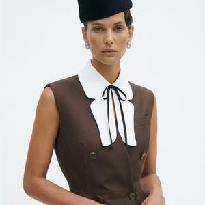 Pu&ograve; includere: Una donna indossa un cappello nero e un gilet marrone con bottoni dorati. Indossa un colletto bianco con una cravatta nera. Il gilet ha un design doppiopetto. Sono visibili anche orecchini d'argento e un anello grande.