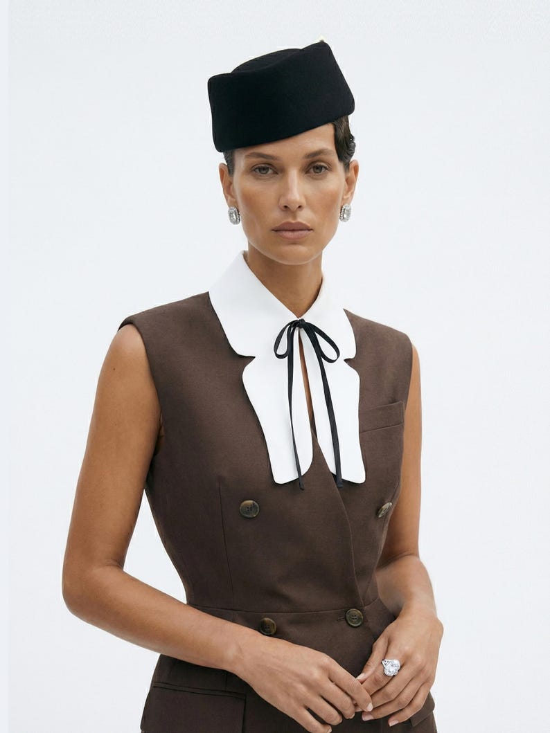 Pu&ograve; includere: Una donna indossa un cappello nero e un gilet marrone con bottoni dorati. Indossa un colletto bianco con una cravatta nera. Il gilet ha un design doppiopetto. Sono visibili anche orecchini d'argento e un anello grande.