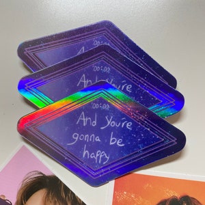 May include: A holographic sticker with a diamond shape and a purple background. The sticker has a white border and the text "And you're gonna be happy" written in white.