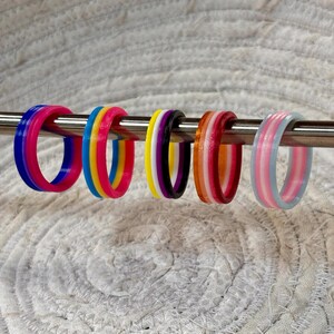 May include: Five circular rings are displayed on a horizontal metal rod. Each ring features distinct color stripes: blue and pink, blue, yellow and pink, yellow, white, black and purple, orange, white and red, and light blue, pink and white.