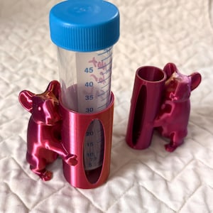 Op de afbeelding: Een magenta testbuishouder met een Frans bulldog ontwerp. De houder heeft een cilindrische vorm met een opening om de metingen op de testbuis te bekijken. Een testbuis met blauwe dop is erin geplaatst.