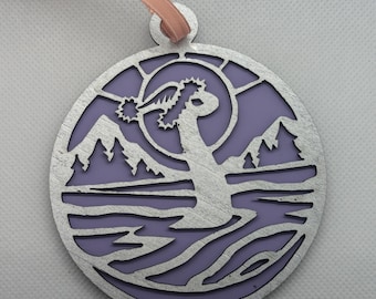 Puede incluir: Adorno redondo plateado con fondo morado y diseño de paisaje. El diseño presenta montañas, un árbol y agua. Se adjunta una cinta rosa claro para colgar.