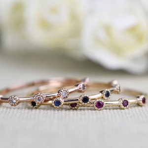 May include: Three delicate gold rings with small gemstones. The top ring has clear stones, the middle ring has blue stones, and the bottom ring has red stones.