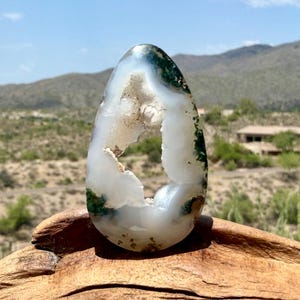May include: A teardrop-shaped moss agate stone featuring a translucent white body with a central hollow geode cavity and deep green moss-like inclusions throughout the crystalline structure, resting on a textured piece of natural brown wood.