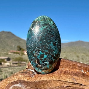 Pode incluir: Uma pedra preciosa polida de formato oval com uma mistura vibrante de azul-petróleo profundo, turquesa brilhante e padrões de matriz de castanho escuro a preto. A pedra tem uma superfície lisa e reflexiva que capta a luz e exibe inclusões.