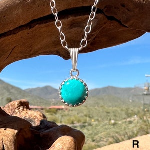 Small round turquoise pendant set in sterling silver, on a 2.7 mm oval link chain, hanging from a piece of wood with the Arizona desert and sky in the background. The gemstone is rich sky blues with faint mottling.
