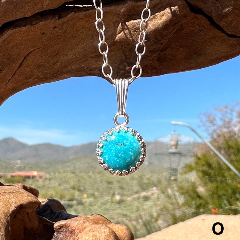 Small round turquoise pendant set in sterling silver, on a 2.7 mm oval link chain, hanging from a piece of wood with the Arizona desert and sky in the background. The gemstone is mottled light and medium sky blues with faint black veining.