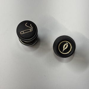 May include: Two black car cigarette lighter plugs. One plug has a white graphic of a lit cigar. The other plug has a white graphic of a flame.