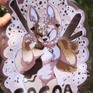 May include: A clear plastic badge holder with a brown and white cartoon animal with the word "COCOA" printed in black at the bottom. The animal has a brown and white spotted pattern and is wearing a collar with a bell.