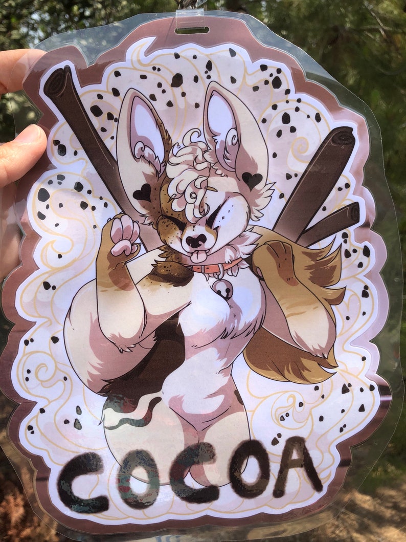 May include: A clear plastic badge holder with a brown and white cartoon animal with the word "COCOA" printed in black at the bottom. The animal has a brown and white spotted pattern and is wearing a collar with a bell.