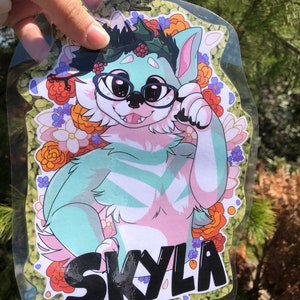 May include: A green and pink furry character with a black name tag that says "Skyla". The character is wearing glasses and a wreath of green leaves and red berries. The character is surrounded by a floral pattern of orange, red, white, and purple flowers.