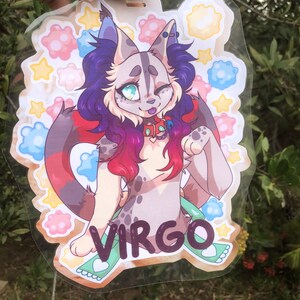 May include: A colorful illustrated badge featuring a cartoon cat with purple and red hair, blue eyes, and a pink nose. The cat is wearing a red collar with a silver charm and a green and white ribbon. The badge has a white background with a variety of colorful flowers and stars. The word "VIRGO" is written in black letters on the bottom of the badge.