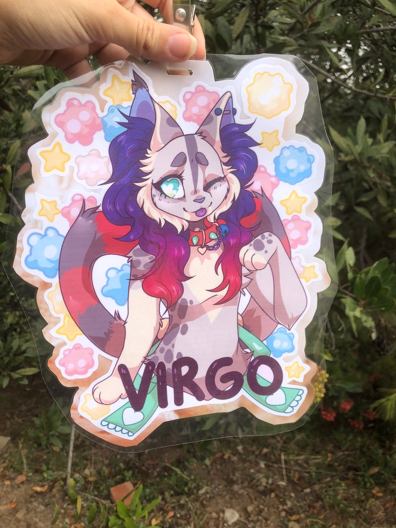 May include: A colorful illustrated badge featuring a cartoon cat with purple and red hair, blue eyes, and a pink nose. The cat is wearing a red collar with a silver charm and a green and white ribbon. The badge has a white background with a variety of colorful flowers and stars. The word "VIRGO" is written in black letters on the bottom of the badge.