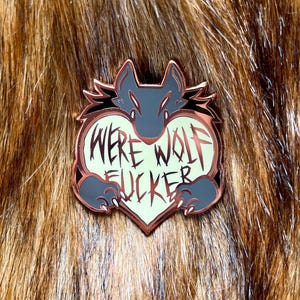 May include: A heart-shaped enamel pin featuring a werewolf head inside. The pin is gold with a black outline and reads "Werewolf Fucker" in a gothic font.