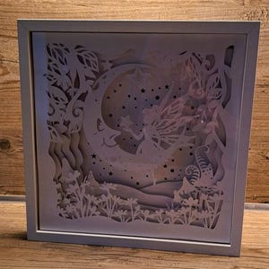 Könnte beinhalten: Eine weiße, gerahmte Schattenbox mit einem 3D-Papier-Ausschnitt-Design einer Fee, die über einen Halbmond mit Sternen fliegt. Die Fee hält einen Stern in ihrer Hand. Das Design ist vor einem Hintergrund aus Blumen und Blättern angeordnet.