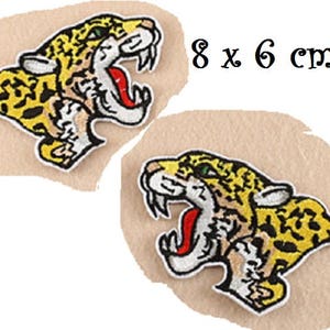 Può includere: Due toppe ricamate raffiguranti una testa di giaguaro ruggente, con dettagli gialli, neri, bianchi e rossi. Ogni toppa misura circa 8 x 6 cm. Il testo "8 x 6 cm" è visibile.