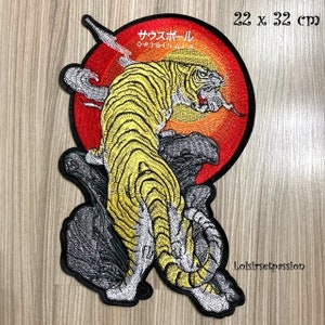 Può includere: Toppa ricamata raffigurante una tigre gialla con strisce nere, ambientata su un sole rosso e arancione. La toppa include testo giapponese e misura 22 x 32 cm. La tigre è appollaiata su una roccia grigia strutturata.
