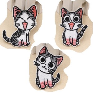Peut inclure: Trois écussons brodés représentant des chats de dessin animé. Chaque chat a un corps blanc, des rayures grises et des détails roses sur les oreilles et les pattes. Les chats affichent différentes expressions, dont un sourire, une surprise et un regard espiègle.