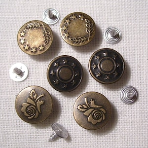May include: An assortment of antique brass-colored jean buttons with various designs, including stars, laurel wreaths, and roses. Each button is round and has a matching silver rivet for attachment. The buttons are arranged on a white fabric surface.
