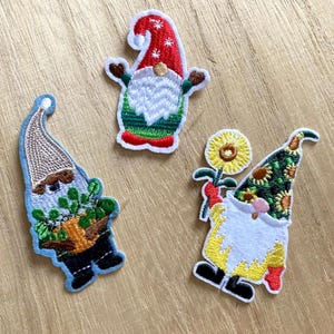 Op de afbeelding: Drie geborduurde strijkpatches met gnomen. Eén gnome draagt een rode muts en houdt een groene plant vast. Een andere gnome draagt een gele muts en houdt een zonnebloem vast. De derde gnome draagt een bruine muts en houdt een groene plant vast.