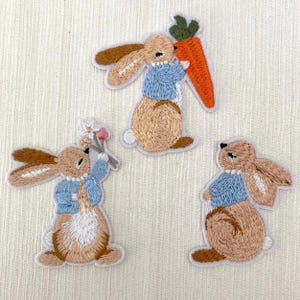Puede incluir: Tres parches de planchado bordados con conejos marrones que llevan camisas azules. Un conejo sostiene una zanahoria, otro una flor y el último está de pie con las patas juntas.