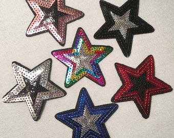 May include: Six sparkly star-shaped iron-on patches in various colors, including pink, black, rainbow, silver, red, and blue. Each star has a silver center and is made of sequins.