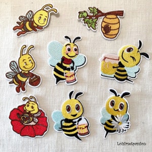 Könnte beinhalten: Eine Sammlung gestickter Bienenaufnäher in verschiedenen Posen. Die Bienen sind gelb und schwarz mit blauen Flügeln, einige halten Honigtöpfe oder Blumen. Ein Aufnäher zeigt einen Bienenstock auf einem Ast. Die Aufnäher befinden sich auf hellem Stoff.